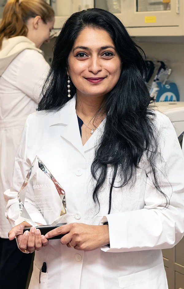 PCOM faculty member Minal Mulye, PhD, smiles in a research laboratory while holding her glass 2025 Northrup Educator Award