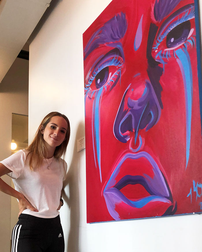 Clinical psychology student Kaitlyn O’Neill (PsyD ’25) smiles as she poses in front of her piece "maybe this is where it ends" in an art gallery