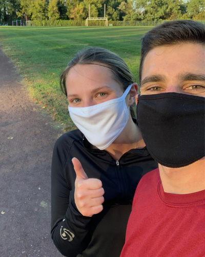 Two PCOM med students wear masks and give a thumbs up in front of a track after completing their individual virtual 5K run as part of PCOM's SOMA fundraising event.