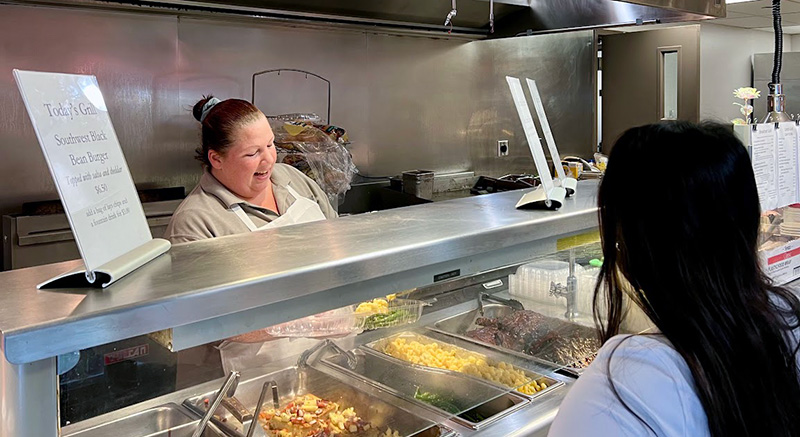 Chef Heather Donahue smiling and serving customers behind the hot food bar in PCOM's cafeteria