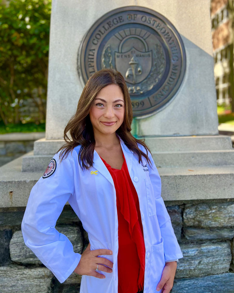 Karli Meller (DO ’26) poses in her medical student white coat next to a pillar with the PCOM crest
