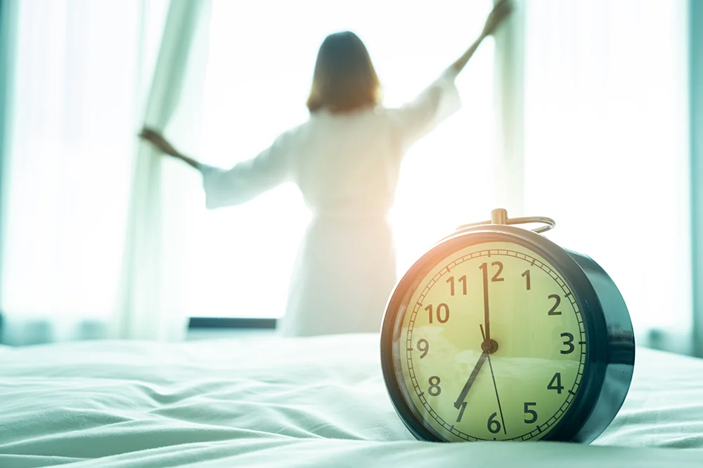 Person in a robe waking up to open the curtains for sunlight with a classic analog alarm clock in the foreground representing daylight savings time sleep adjustments