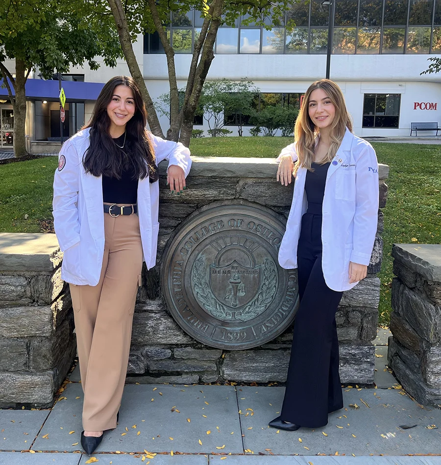 Med students and sisters Kaylyn Branton (DO '28) and Megan Branton (DO '29) pose against the PCOM college seal in the courtyard wearing their white coats