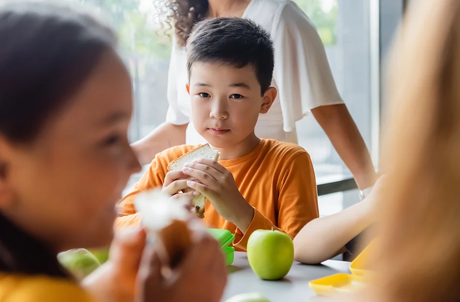 A child holding a sandwich while sitting on a table in a school cafeteria