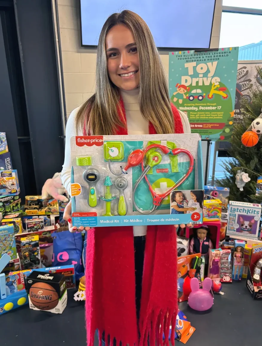 PCOM medical student Ellie Koerner (DO '29) smiles while holding a gift in front of a pile of toys during her toy drive event