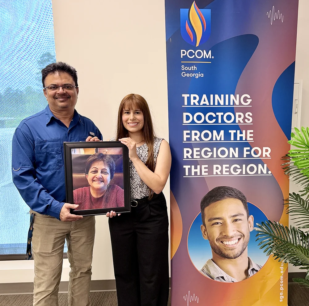 Shiv Dhiman, MD, and Selena Meza (CGS '26, DO '30) holding a picture of the late Savita Arya, MD, inside a PCOM South Georgia classroom