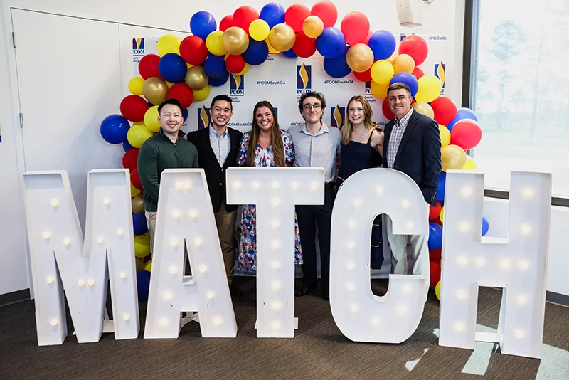 DO students in formal wear pose under an arch of balloons and behind a giant "MATCH" sign
