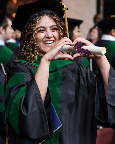 PCOM South Georgia graduate smiles on stage and makes heart symbol with hands with holding diploma