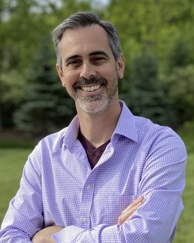 Headshot of Alexander Ophir, PhD, smiling and dressed in a purple dress shirt with green foliage in background