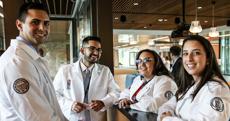 Four PCOM South Georgia medical students smiling in the lobby wearing their physician white coats.