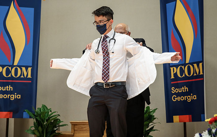 A member of the DO Class of 2025 puts on his white coat with help from a faculty member