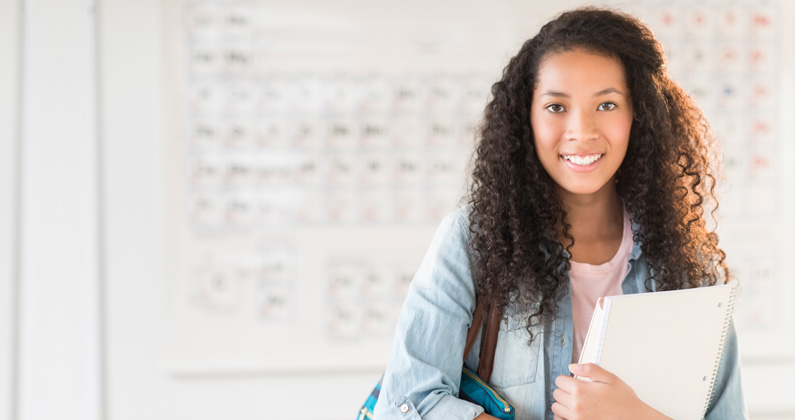 High school student holds books and smiles in front of a whiteboard with science lessons