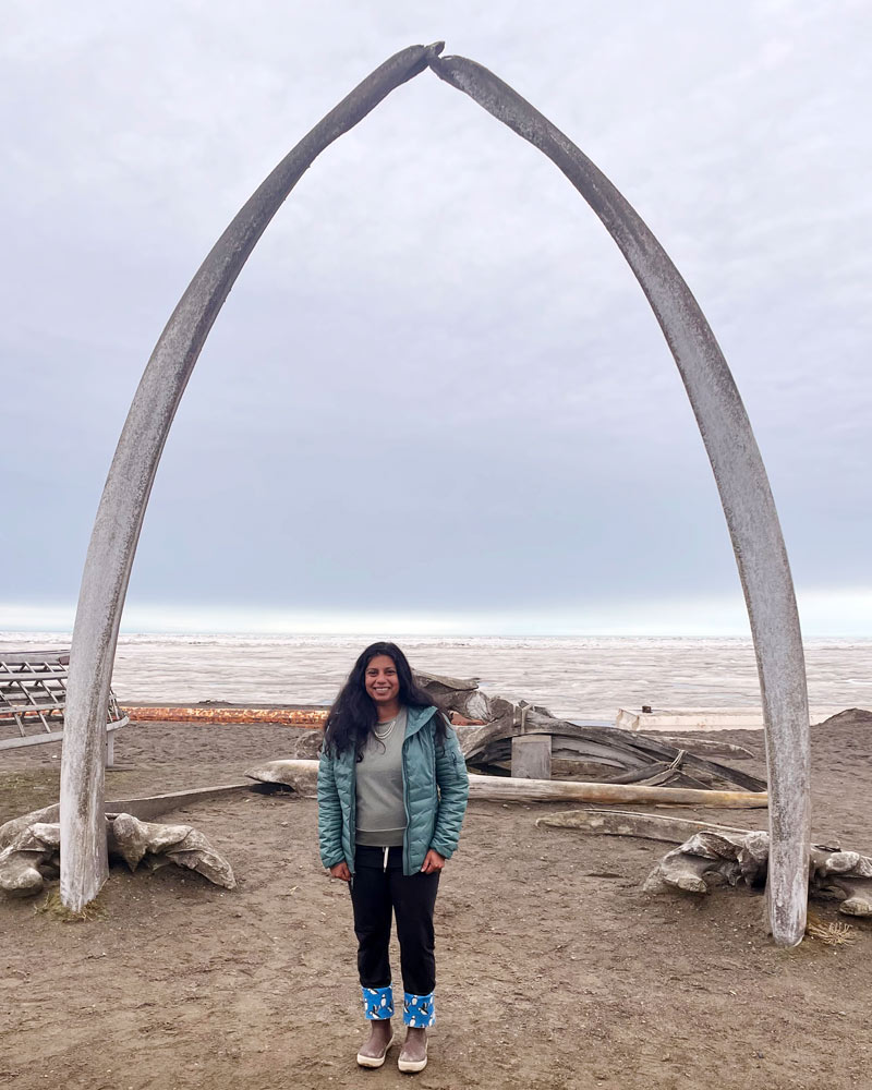 Nilam Vaughan, DO ’11, poses for a photo on a beach beneath an archway made from whale bones while on assignment in Utqiagvik, Alaska