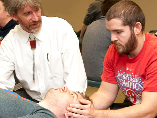 Professor instructs a student 1-on-1 during an OMM lab session while he manipulates a student's head