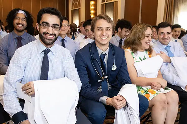 PCOM DO students sit in a row and smile as they hold their white coats during the White Coat Ceremony