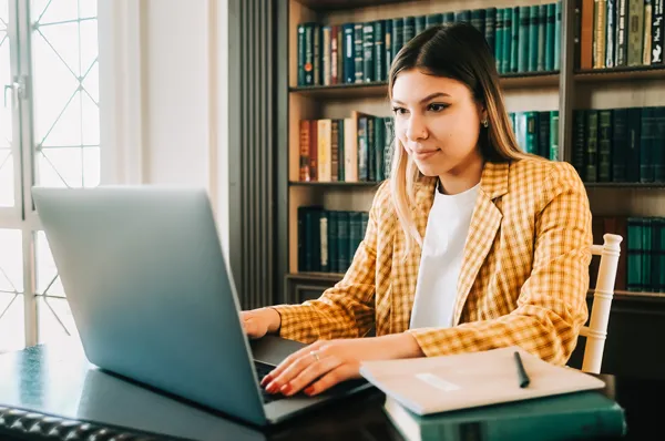 A college student using a laptop in a library while attending PCOM's School Psychology Virtual Information Session