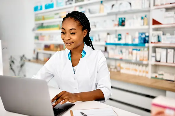 Pharmacist in white lab coat stands at drugstore counter and works on laptop computer