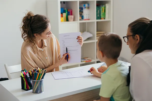 A woman seated at a desk holds up a piece of paper and points out text for a mother and son