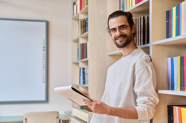 Man leans against bookshelf while reviewing note pad
