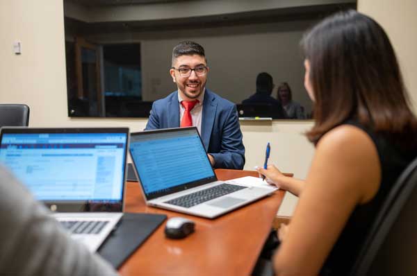 A smiling male pharmacy student wearing a suit is sitting at a table speaking with two other people using computers.