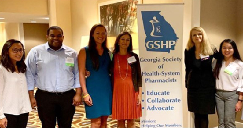 PCOM Georgia pharmacy students and faculty smile in front of the GSHP Summer Meeting conference sign.