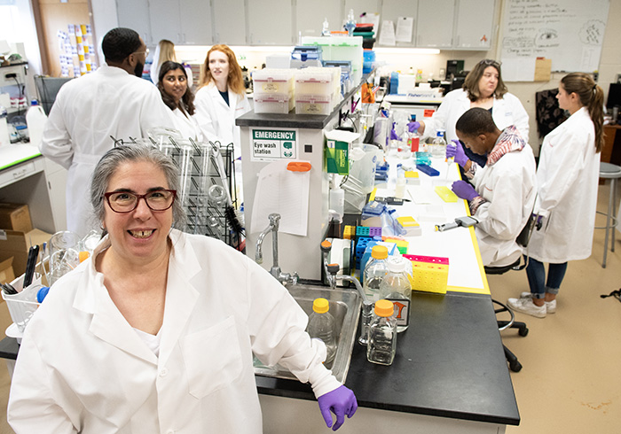 Marina D'Angelo smiles in her research white coat while her osteoarthritis research team works in a PCOM laboratory