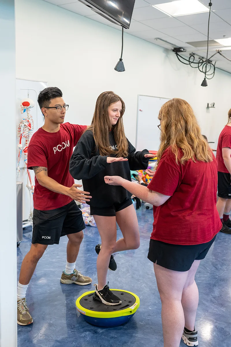 Vertical shot of DPT students smiling and helping one another with a balance exam exercise