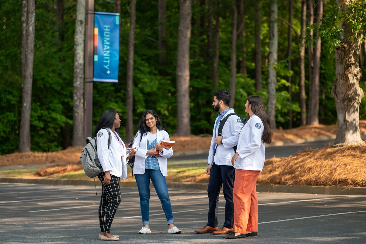 Students smiling and talking with backpacks in the parking lot of PCOM Georgia with pine trees in the background