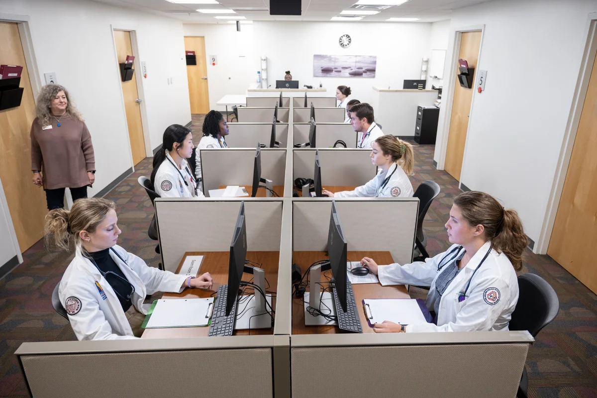 PCOM Georgia students in white coats smile as they use computers in the Sim Center exam debriefing area