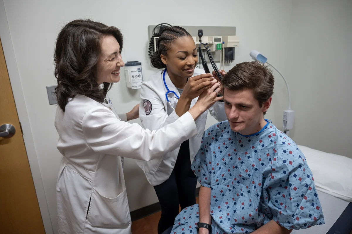 PCOM faculty member and med student smile as they exam the ears of a patient actor in the Sim Center