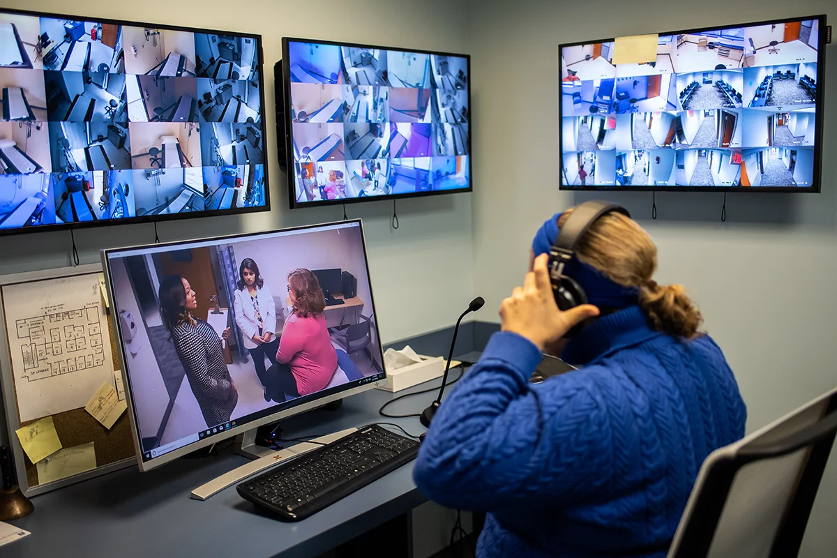 A PCOM staff member puts on a pair of headphones as she looks at computer monitors and equipment within the Simulation Center's Control Room