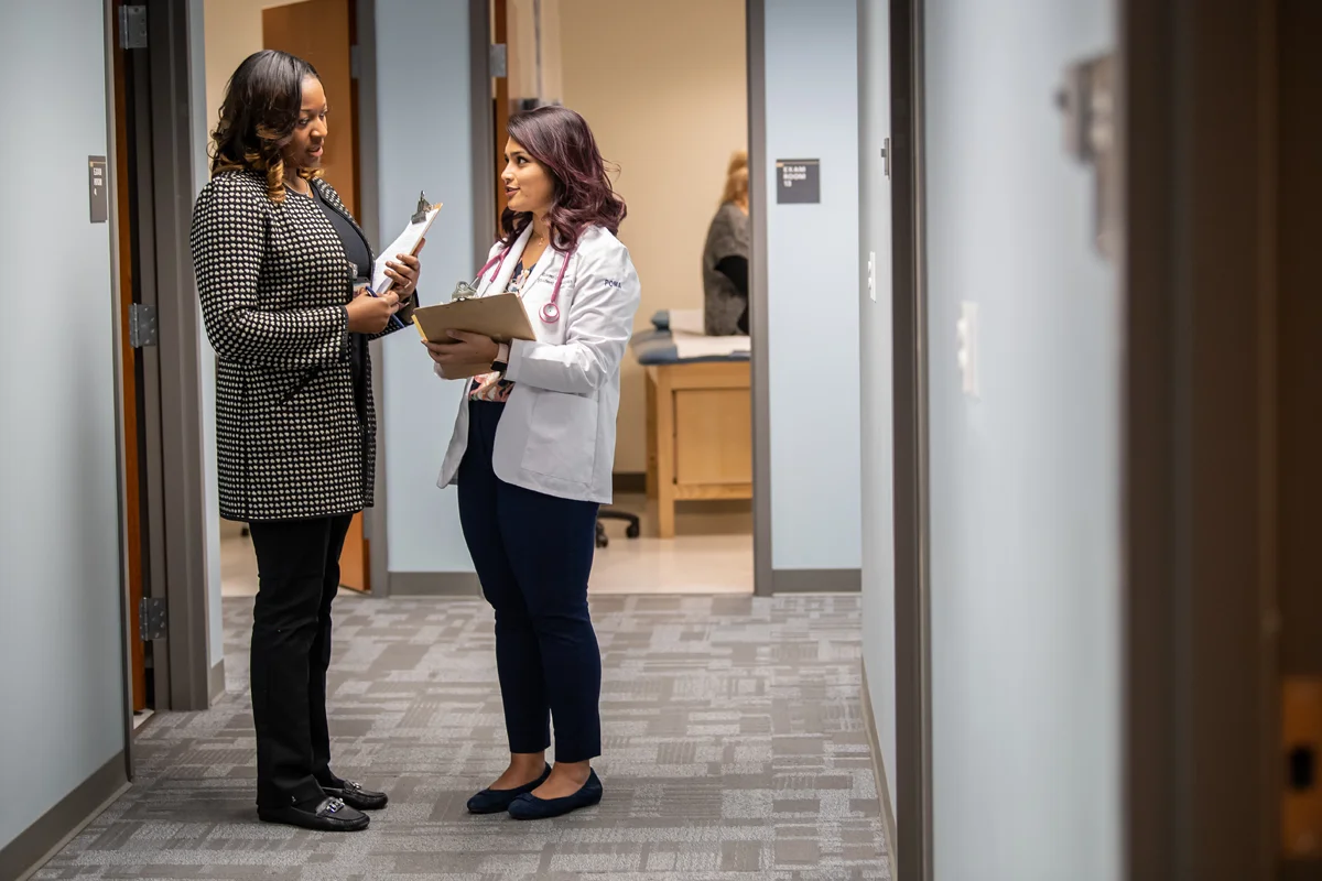 Two students talking in a hallway in the CLAC. A standardized patient can be seen in the exam room in the background.