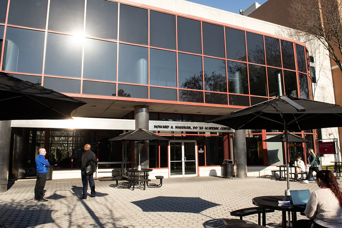 Students studying and staff talking on tables in the courtyard front of Hassman Academic Center