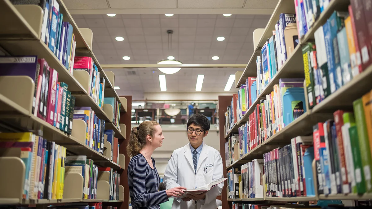 PCOM medical students looking at a book within bookshelves in the library