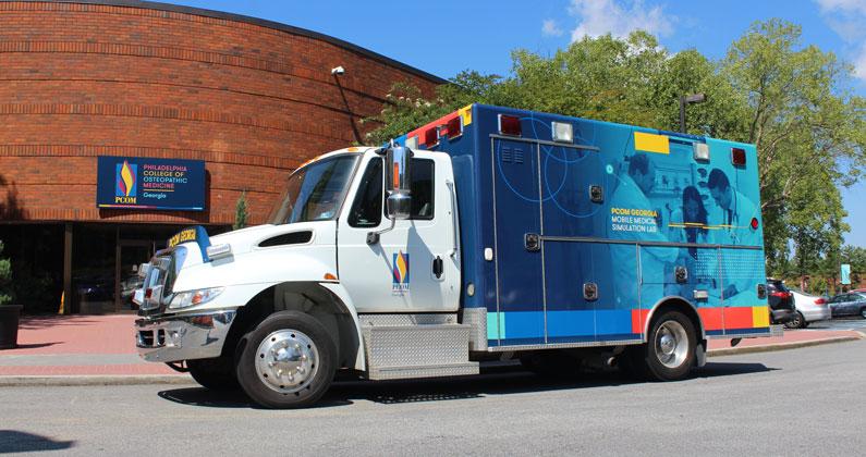 A heavy-duty mobile medical laboratory with PCOM branding wrapped around the rear half of the vehicle is parked in front of the PCOM Georgia campus.