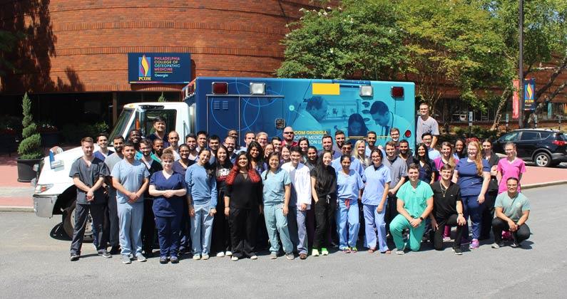 A group of students and staff dressed in hospital scrubs and white lab coats pose outside the PCOM Georgia campus front entrance with the new mobile medical laboratory vehicle.