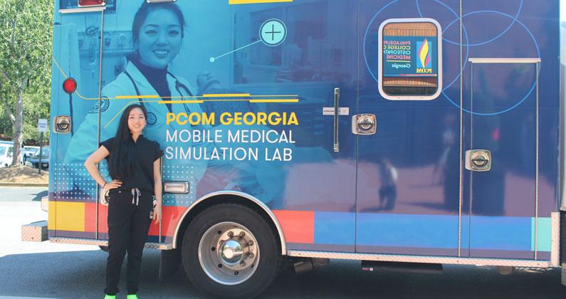 A medical student stands beside the mobile medical laboratory. A photo of the student in a white lab coat is used as part of the PCOM branding wrapped around the vehicle.