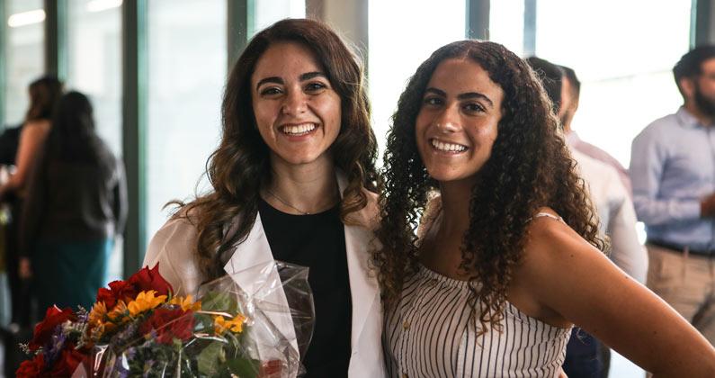 Two students pose for the camera. One is wearing a white lab coat and is holding a bouquet of flowers.