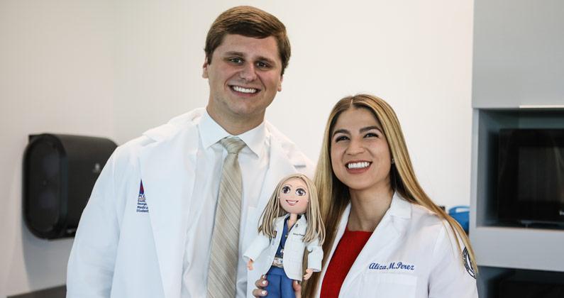 Two medical students pose together wearing their white coats. One is holding a small doll wearing a white coat. The doll resembles the student.