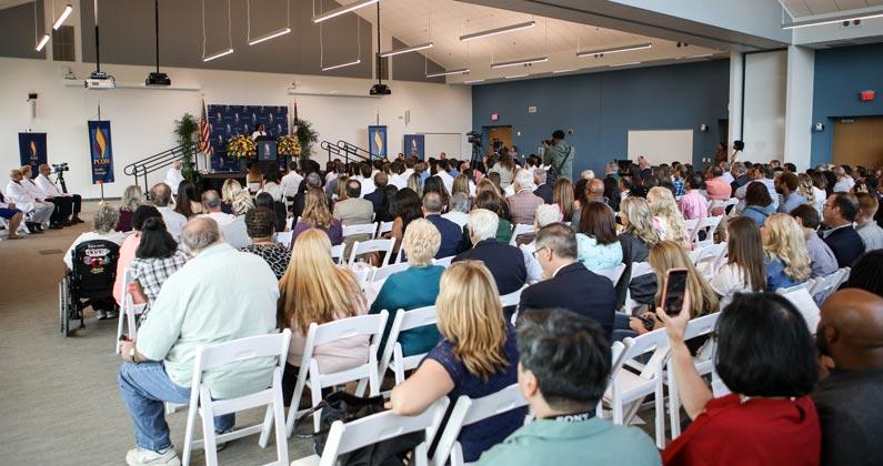 An audience is seated in white folding chairs in a large conference room, their attention focused on the stage at the front of the room.