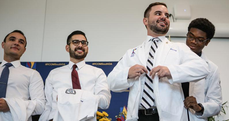 Medical students stand on stage waiting to receive their white coats. One student has just received their coat and is wearing it.