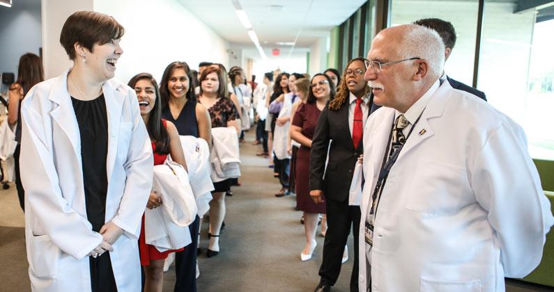 Medical students form two single-file lines behind faculty members in a long hallway.