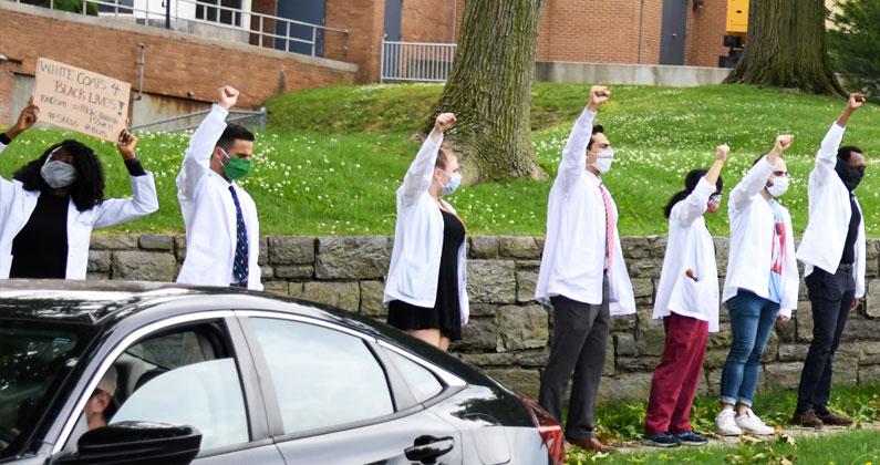 PCOM medical students wear their physician white coats, stand along the side of City Ave and raise their fists in the air.