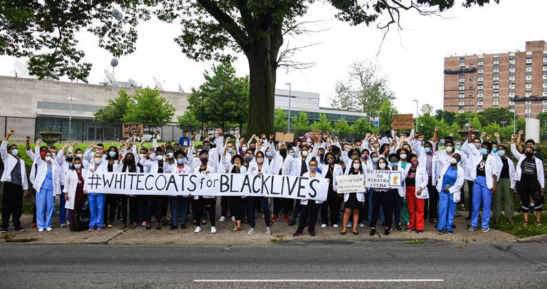 Dozens of PCOM students, faculty and staff stand for a group photo next to City Ave and hold a 'White Coats for Black Lives' banner.