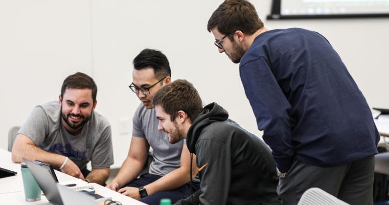 Four male medical students gather and smile over a laptop in a PCOM South Georgia classroom during the winter term.