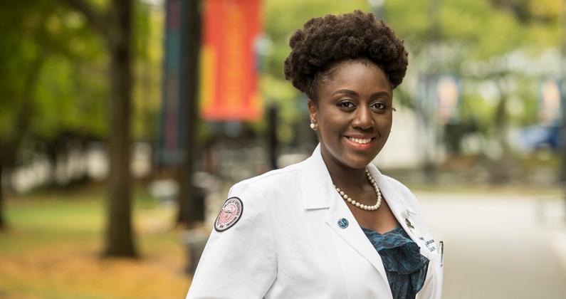A female DO medical student smiles while wearing her physician white coat on a walkway