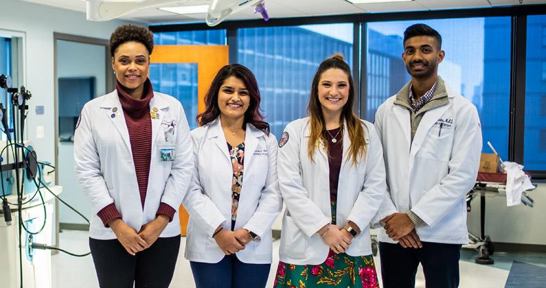 Diverse group of four DO medical students wearing physician white coats smile in front of medical simulation equipment at PCOM