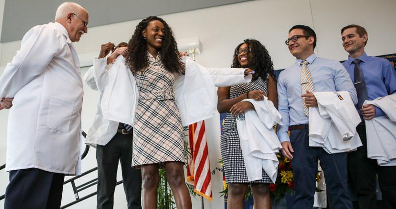 A Black female medical student smiles as a faculty member places the physician white coat on her shoulders during the ceremony at PCOM South Georgia