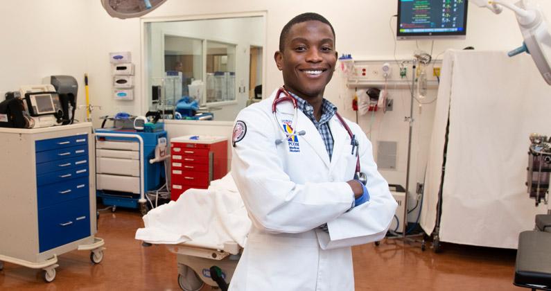 A Black male medical student poses in his physician white coat in front of a surgery table at the PCOM Georgia simulation suite