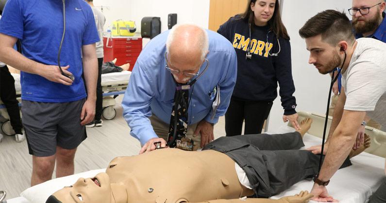 Dr. Fredrick demonstrates how to use a stethoscope on a simulation mannequin while DO med students watch.